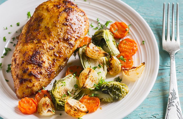 baked chicken breast with brussels sprouts, onions and carrots on a white plate on wooden surface